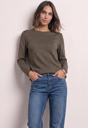 Kvinde med skulderlangt brunt hår iført olivengrøn langærmet skjorte og blå jeans, hænderne i lommerne, står foran en ensfarvet lys baggrund.