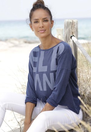 Long-sleeve navy blue top with bold gray letters, paired with white skinny jeans. The top features a relaxed fit and soft texture.