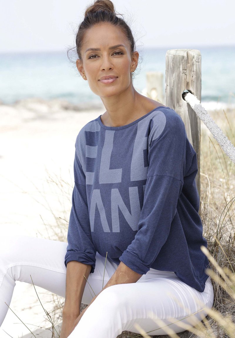 Langärmliges, marineblaues Oberteil mit kräftigen grauen Buchstaben, kombiniert mit weißen Skinny-Jeans. Das Oberteil hat eine lockere Passform und eine weiche Textur.
