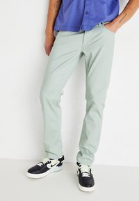 Pantalones ajustados de color verde claro, hechos de una mezcla de algodón, con piernas rectas. Llevados con zapatillas negras y blancas y una camisa de manga corta azul.