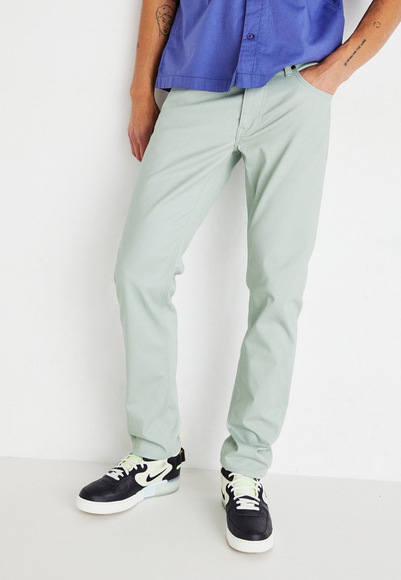 Pantalones ajustados de color verde claro, hechos de una mezcla de algodón, con piernas rectas. Llevados con zapatillas negras y blancas y una camisa de manga corta azul.