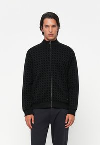 Zwarte zip-up jas met een patroontextuur. Heeft een geribbelde kraag, manchetten en zoom. Zijstrepen voegen detail toe. Gladde stofafwerking.
