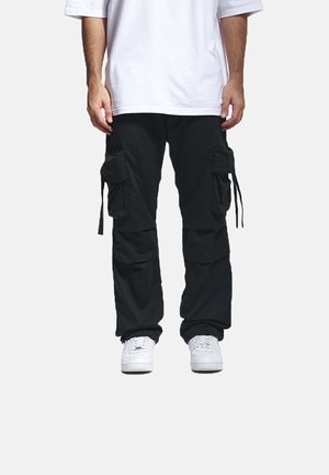 Svarte cargo-bukser med flere sidelommer, justerbare stropper og en avslappet passform, kombinert med hvite joggesko og en løs hvit t-skjorte.