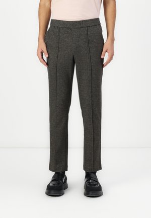Mann trägt maßgeschneiderte dunkelgraue Hose mit Frontfalten und schwarzen Lederschuhen, steht vor weißem Hintergrund.