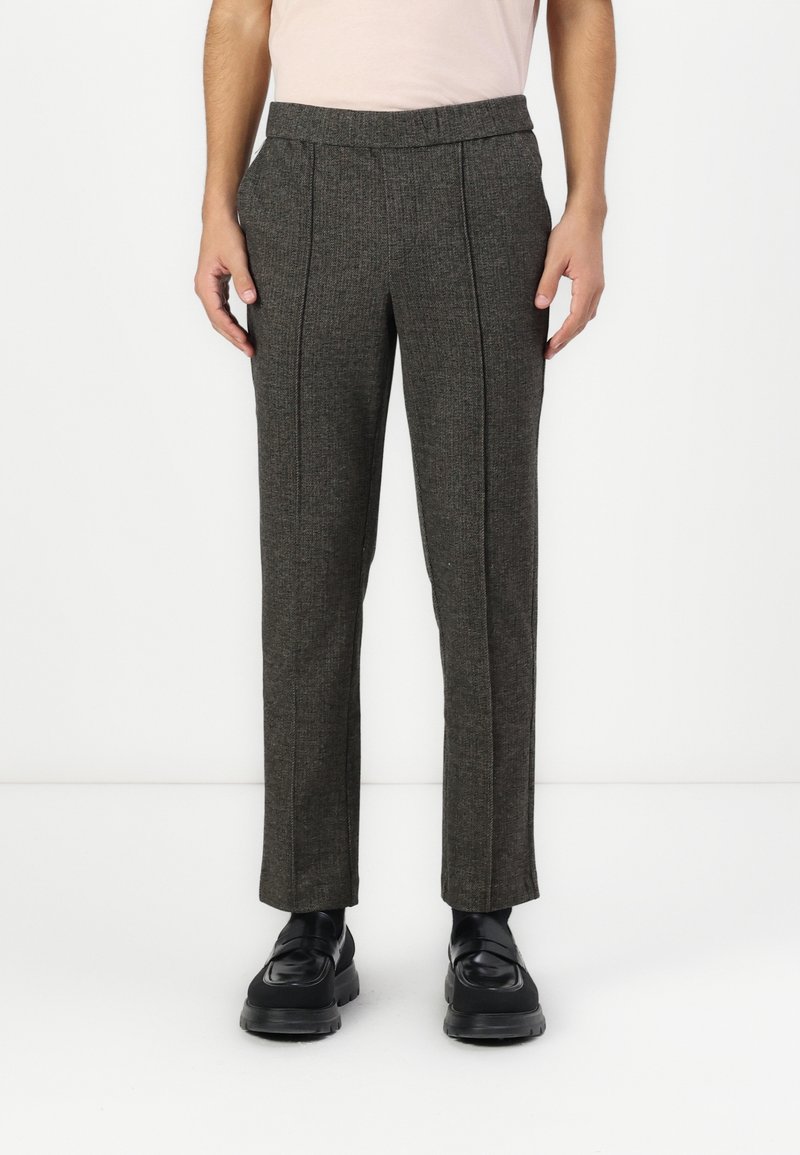 Homme portant un pantalon gris foncé sur mesure avec plis à l'avant et des chaussures en cuir noir, debout sur un fond blanc.