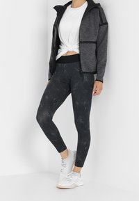 Leggings sportive neri con una finitura testurizzata, abbinati a una t-shirt bianca annodata e a una giacca grigia con zip e accenti a contrasto.