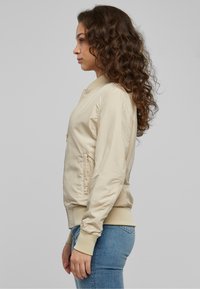 Veste bomber beige à finition lisse, avec poignets et ourlet côtelés, poches latérales et fermeture éclair, portée avec un jean bleu ajusté.