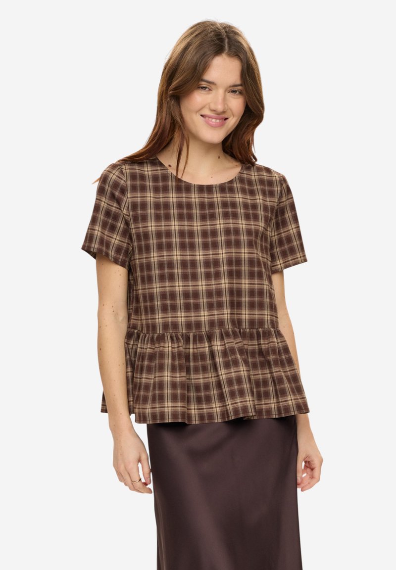 Jeune femme souriante, portant un haut à manches courtes à carreaux marron et beige avec un ourlet à volants, et une jupe marron foncé.