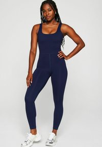 Navy blue athletic jumpsuit with a square neckline, fitted design, and seamless texture, complemented by silver sneakers.
