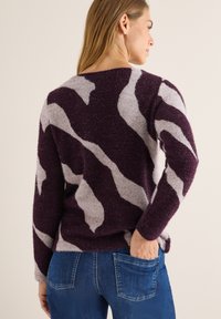 Fioletowy i szary sweter w wzór chevron z miękkiego, teksturowanego materiału. Długie rękawy i luźny krój, zestawiony z niebieskimi dżinsami.