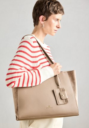 Beige draagtas met een gestructureerde afwerking, voorzien van een voorzak en gouden hardware. De tas wordt gecombineerd met een gestreept trui.