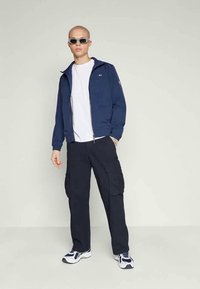 Granatowa kurtka zip-up z stójką, biała koszulka, ciemne spodnie cargo z kieszeniami oraz buty sportowe, model w okularach przeciwsłonecznych.
