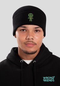 Giovane uomo che indossa un berretto di lana nero con un patch di un pupazzo ricamato verde e una felpa nera con la scritta "WRONG FRIENDS" in turchese.