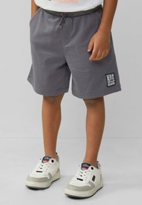 Grijze katoenen shorts met een elastische tailleband en trekkoord, met een klein logopatch aan de linkerkant. Gecombineerd met witte sneakers.