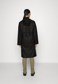 Gabardina negra hasta la rodilla con capucha, que presenta un cintura con cordón y mangas largas. Llevada sobre pantalones beige y zapatos negros.