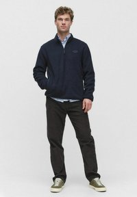 Granatowy polar z zamkiem błyskawicznym i bocznymi kieszeniami, w połączeniu z czarnymi spodniami i zielonymi trampkami. Charakteryzuje się gładką teksturą i casualowym stylem.