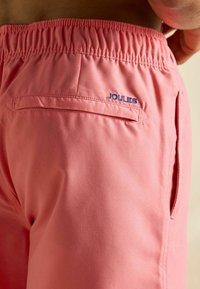 Ružové šortky s elastickým pásom, obsahujú bočnú kapsu a skrytú zadnú kapsu. Značka "JOULES" je vyšitá modrou farbou.