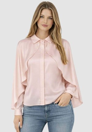 Donna con lunghi capelli biondi che indossa una camicetta di raso rosa chiaro con maniche stile mantella e jeans blu, in piedi davanti a uno sfondo semplice.