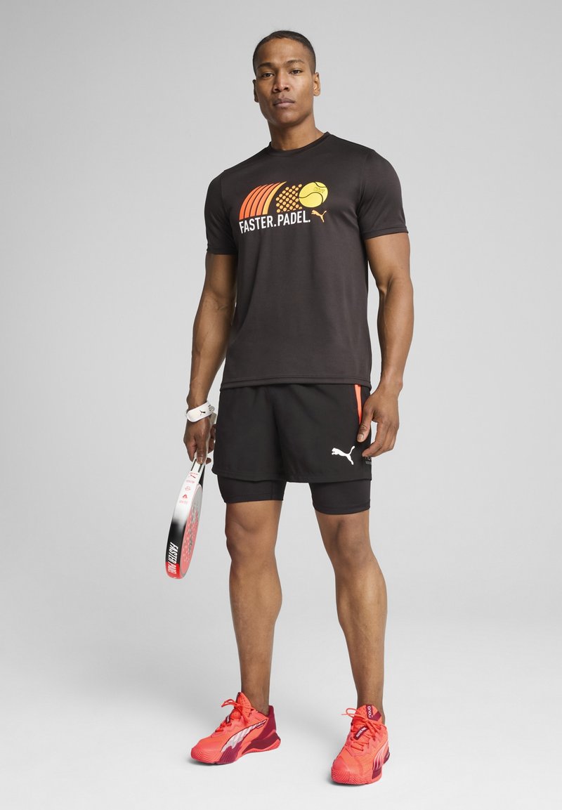 Schwarzes Sport-T-Shirt mit orangefarbenem und gelbem grafischem Text "FASTER.PADEL", kombiniert mit schwarzen Shorts. Zubehör umfasst ein weißes Handgelenkband und einen Schläger mit roten Akzenten.