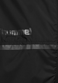 Zwarte jas van een gestructureerde stof, met een reflecterende strook en een zilveren "hummel" logo. Glad oppervlak met een functioneel ontwerp.