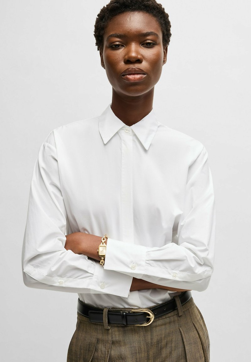 Camisa blanca de manga larga con cuello clásico, combinada con pantalones de cuadros y una pulsera dorada que realza el look. Textura de tela suave.