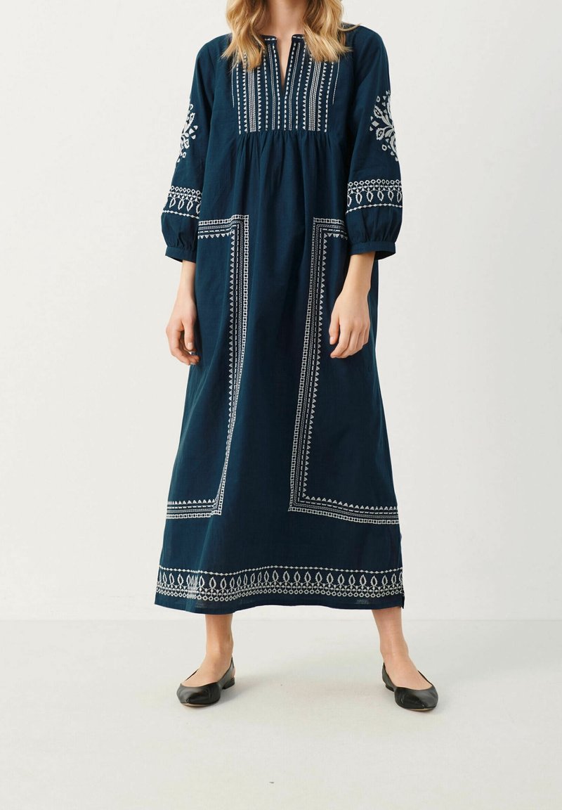 Femme portant une longue robe bleu marine avec une broderie géométrique blanche, manches trois-quarts et chaussures plates noires, debout devant un fond clair uni.