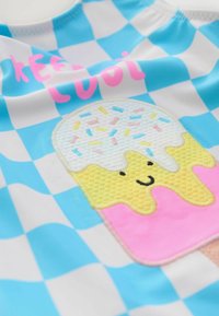 Maillot de bain avec un motif à carreaux bleu et blanc, orné d'un graphique de cône de glace texturé en jaune et rose, accompagné de la phrase "restez au frais" en rose.