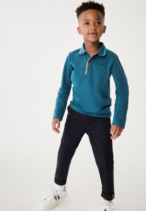 Teal langærmet polo med en knappet halsudskæring, kontrastfarvet krave og broderet logo, som er parret med mørke bukser og hvide sneakers.