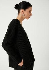 Mustat ribattuja neulepusero V-puoleisella kaula-aukolla, pitkillä hihoilla ja sivurei'illä. Mallilla on hopeiset renkaat korvissa. Yksinkertainen, teksturoitu muotoilu.