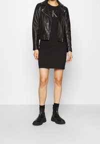 Czarna skórzana kurtka motocyklowa z asymetrycznymi zamkami błyskawicznymi, zestawiona z dopasowaną czarną sukienką i masywnymi czarnymi botkami za kostkę. Prosty fason, gładka faktura.