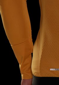 Yellow athletic long sleeve top with textured fabric, featuring a sleek design and reflective accents near the hem. Black waistband at the bottom.