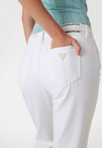 I jeans in denim bianco presentano un design aderente con una tasca posteriore cucita, che mostra un piccolo logo triangolare dorato. Il tessuto appare liscio e pulito.