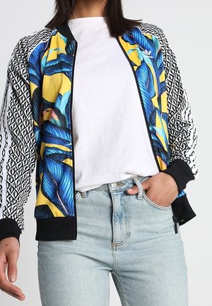 Vrouw draagt een kleurrijk, bloemenpatroon jas met zwart-witte geometrische mouwen, gecombineerd met een wit t-shirt en lichtblauwe jeans.