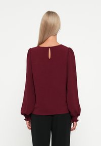 Bordeauxbluse med lange, puffede ærmer, tekstureret stof og en lille nøglehulåbning ved nakken bagpå. Kombineret med sorte bukser.