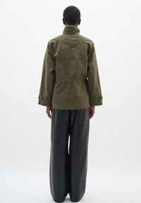 Olivgrön jacka med hög krage, dragsko i midjan och knäppta ärmar, kombinerad med svarta läderbyxor med vida ben. Minimalistisk design.