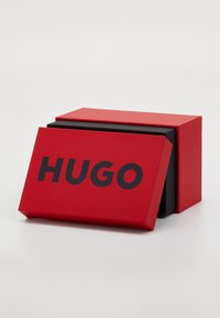 Punane ristkülikukujuline kast, millel on must sisemine osa, esiküljel on paksu musta fondiga "HUGO". Sile, mati tekstuur.