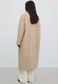 Žena s dlouhými rovnými vlasy oblečená v dlouhém béžovém vlněném kabátu, krémových širokých kalhotách a tmavých špičatých botách stojí u prosté stěny.