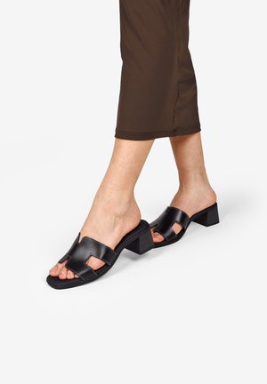 Mules de tacón bloque negro con un diseño recortado en la parte superior. Material de cuero suave con detalles de costura en contraste.