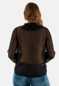 Blusa nera trasparente con maniche a sbuffo lunghe, colletto arricciato, tessuto fluido e vita aderente. Vista da dietro.