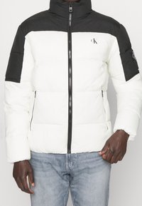 Mężczyzna w białej i czarnej kurtce puchowej z przednim zamkiem błyskawicznym i bocznymi kieszeniami, w połączeniu z jasnoniebieskimi jeansami.