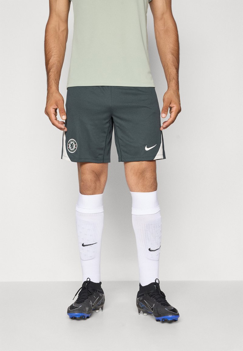 Gray athletic shorts with Chelsea logo; white Nike swoosh accent. Model wears white socks and black soccer cleats with blue details.