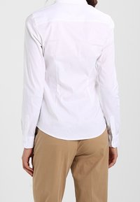 Weißes Langarmhemd mit spitzem Kragen, geknöpften Manschetten und abgerundetem Saum. Der Stoff wirkt glatt mit minimaler Textur.