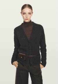 Ciemnoszara sweter z warkoczykiem i dekoltem w serek, z teksturą ściągacza oraz przednimi guzikami. Połączony z brązowym golfem i ciemnymi spodniami, z brązową torbą na pasku.