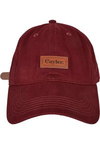 Red cotton cap with a curved brim and a leather patch labeled "Cayler" on the front. Features stitched details and ventilation holes.
