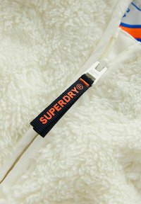 Baltas, pūkuotas fliso audinys su užtrauktuku. Juodame labelyje raudonai oranžinėmis raidėmis parašyta "SUPERDRY®". Užtrauktuko trauklė balta.