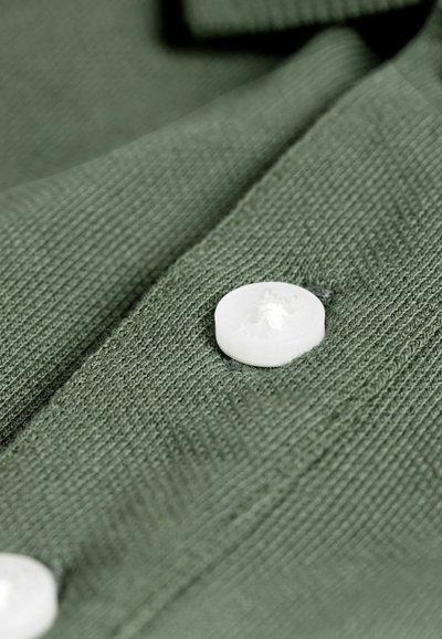 Žalia medžiaga su minimaliai tekstūruota juostele ir ryškiai matoma balta plastikinė saga, demonstruojanti paprastą ir funkcionalų dizainą.