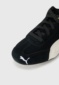 Baskets en suède noir avec des lacets, arborant un logo Puma blanc et des accents beiges sur le côté. Texture lisse et design incurvé.