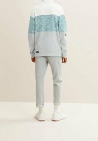 Pullover grigio chiaro con un motivo a righe teal e bianche, abbinato a jeans grigio chiaro e sneakers bianche. Look casual e vestibilità rilassata.