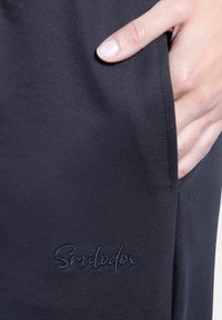 Zwarte joggershort van een zachte, gestructureerde stof, met zijzakken en een geborduurd "Smilodex"-logo op de linker dij.