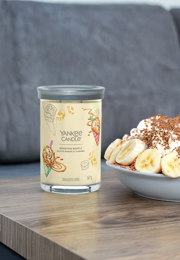 Yankee Candle en un frasco de vidrio transparente con una tapa plateada, con una etiqueta amarilla que presenta ilustraciones de helado y plátanos, colocado al lado de un postre.